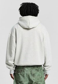 Felpa grigia con cappuccio e vestibilità ampia, caratterizzata da polsini e orlo a coste, indossata con pantaloni verdi a trama e un accento di catena.