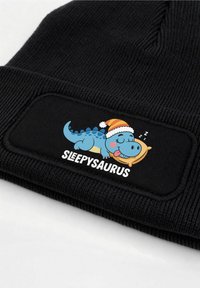 Black ribbed beanie with a fabric patch featuring a blue dinosaur cartoon wearing a red and yellow striped hat, text reads "SLEEPYSAURUS."