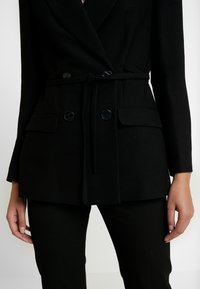 Blazer negro con diseño envolvente, que incluye un cinturón atado, dos bolsillos de parche y botones redondos. Textura suave con hombros estructurados.