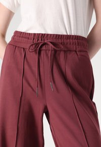 Burgundy jogger pants with an elastic waistband, adjustable drawstring, front pleats, and silver-accented hardware. Soft, textured fabric.