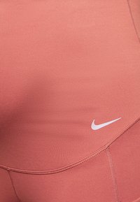 Leggings Nike di colore rosa corallo, realizzati in un tessuto morbido, con un design aderente e un logo swoosh bianco. I dettagli delle cuciture sono visibili vicino alle cuciture.