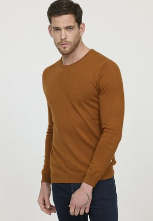 Pull en maille marron avec un col rond et des manches longues. Poignets et ourlet côtelés. Porté avec un jean bleu foncé. Texture lisse, détails minimaux.