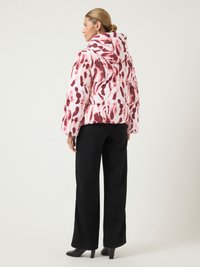 Mulher com penteado em coque, vestindo um casaco acolchoado com capuz de padrão branco e vermelho, calças largas pretas e sapatos de salto pretos, voltada para o lado.