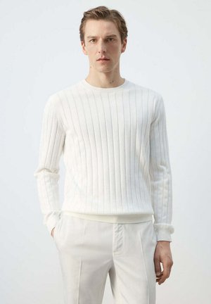 Joven con el pelo corto, vistiendo un jersey blanco de manga larga con canalé y pantalones blancos, de pie con una mano en el bolsillo.