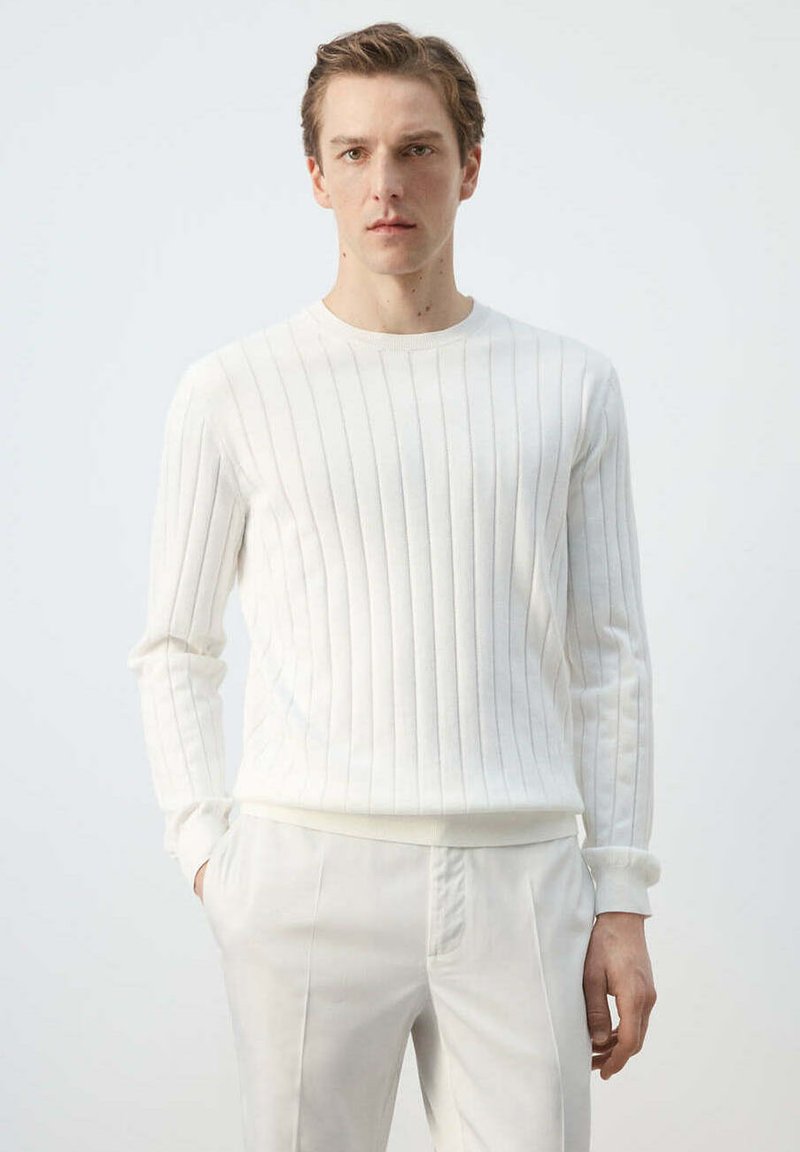 Jeune homme aux cheveux courts portant un pull blanc à côtes à manches longues et un pantalon blanc, debout avec une main dans la poche.