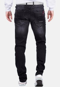 Jeans in denim nero con vestibilità slim, leggera sfumatura, tasche posteriori a toppa e dettagli in cucitura rossa. Indossati con scarpe Converse bianche.