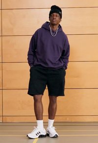 Paarse hoodie, zwarte korte broek, witte sneakers met zwarte accenten. De achtergrond bestaat uit houten panelen en een gymzaal vloer.