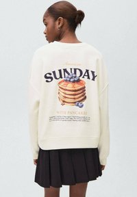 Donna con i capelli raccolti in un bun che indossa un sweatshirts bianco, mostrando una torre di pancakes con mirtilli e il testo "Domenica con pancake" sulla schiena.