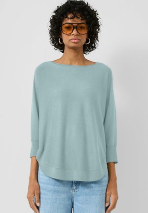 Frau mit dunklen lockigen Haaren, die übergroße bernsteinfarbene Sonnenbrillen, silberne Creolen, einen hellblauen weiten Pullover und hellblaue Jeans trägt.