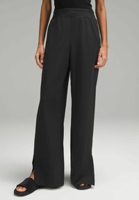 Zwarte wijde broek met een gladde textuur, hoge taille en zijsplitten aan de zoom, gecombineerd met zwarte slidesandalen.