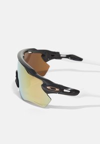 Des lunettes de sport noires et transparentes présentent une lentille surdimensionnée avec une teinte dégradée, accentuée par un logo en or rose et des branches en caoutchouc élégantes.