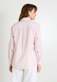 Chemise rayée rose clair à manches longues, avec de petites accents brodés. Les poignets boutonnés et le col classique complètent le design.