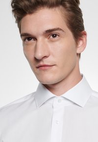 Vit skjorta med knappar och en strukturerad krage. Slätt tyg med en subtil textur. Har en framplacket med två knappar och en klassisk design.