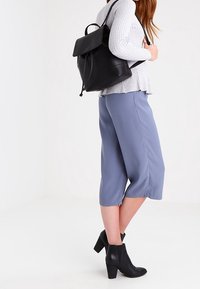 Sac à dos en cuir noir avec une fermeture à rabat, associé à un pull en maille gris clair et un pantalon court bleu. Des bottines noires complètent le look.