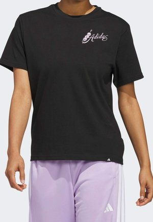 Osoba ubrana w czarną koszulkę Adidas z białą grafiką grilla na patyku i logo w formie pisma ręcznego, do której dopasowane są jasnofioletowe spodnie Adidas z białymi paskami.