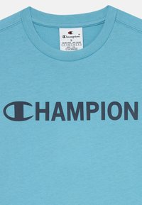 Gaiši zila kokvilnas T-krekls ar apaļu kaklu un uzrakstu "CHAMPION" melnā krāsā, kas izceļas uz krūškurvja, ar emblēmu pa kreisi.