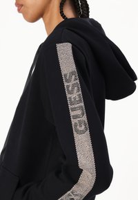 Sudadera con capucha negra con acentos de pedrería a lo largo de la manga, que presenta un bolsillo delantero y una tela suave y texturizada.