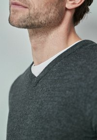 Ciemnoszary sweter z dekoltem w serek, wykonany z miękkiej dzianiny, nałożony na biały t-shirt z okrągłym dekoltem, prezentujący fakturowane wykończenie.