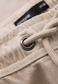 Shorts en tissu beige avec une taille à cordon de serrage. Présente un œillet en métal argenté avec le texte "ombre" et une surface texturée.