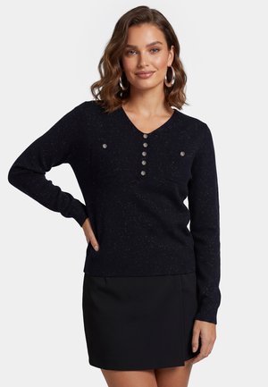 Kvinde med bølget brunt hår iført øreringe, en sort knappet sweater med to brystlommer og et sort skørt, stående med den ene hånd på hoften.