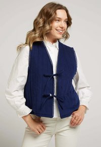 Gilet matelassé bleu marine avec texture côtelée, doté d'une fermeture à crochets et d'un ourlet à pompons sur les bords, porté sur une chemise blanche.