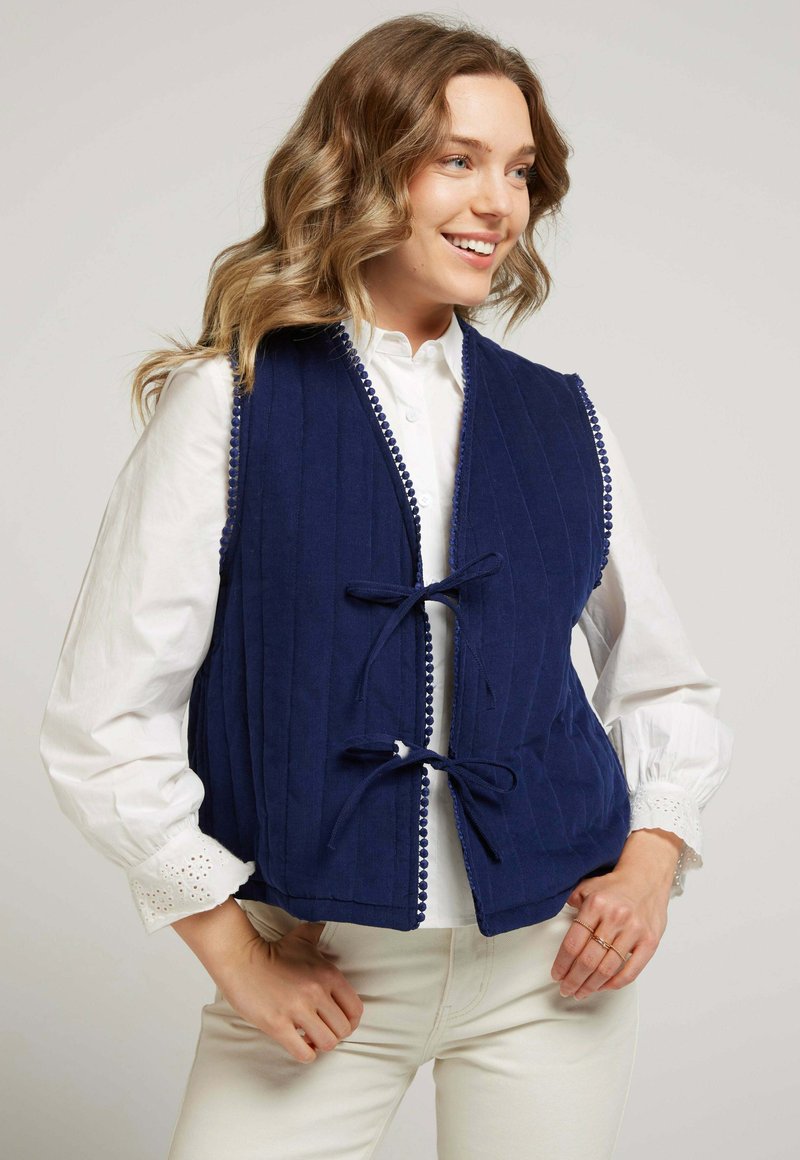 Gilet matelassé bleu marine avec texture côtelée, doté d'une fermeture à crochets et d'un ourlet à pompons sur les bords, porté sur une chemise blanche.