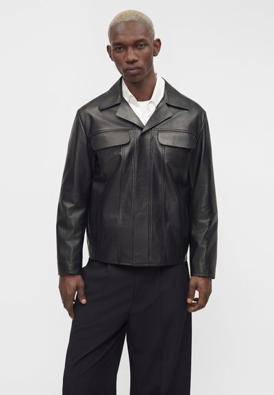 Homme aux cheveux blonds courts portant une veste en cuir noire par-dessus une chemise blanche et un pantalon noir, debout devant un fond clair uni.