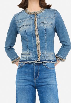 Mujer que lleva una chaqueta vaquera azul ajustada con adornos de cadena dorada en el cuello, los puños y la parte delantera, combinada con vaqueros azules de talle alto.