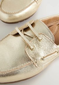 Kulta metallinväriset kengät, joissa on teksturoitu nahkapinta, pyöreä kärkimuoto, nyörityssuunnittelu ja pehmeä beige sisäosa. Varustettu kultaisilla silmukoilla ja ompeleilla.