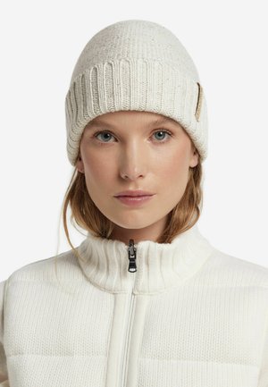 Witte gebreide beanie met een geribbeld ontwerp, textuurpatroon en een klein merkteken aan de zijkant. Draagt het in combinatie met een witte vest met rits.