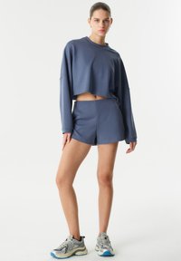 Světle modrý zkrácený sweatshirt a odpovídající šortky vyrobené z měkkého materiálu. Model má na sobě šedé tenisky s modrými a krémovými akcenty.