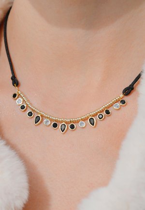 Collier doré avec des pierres noires et transparentes en formes de goutte et ronde, monté sur un cordon noir tressé.