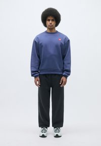 Sudadera púrpura con un logo rojo, combinada con pantalones negros de corte holgado. El modelo tiene un peinado afro y lleva zapatillas negras y blancas.