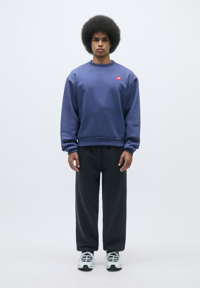 Sudadera púrpura con un logo rojo, combinada con pantalones negros de corte holgado. El modelo tiene un peinado afro y lleva zapatillas negras y blancas.