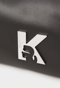 Juodas tekstūruotas odos krepšys su ryškiu sidabriniu "K" logotipu ir silueto detale. Tvarkingas, modernus dizainas su blizgiais akcentais.