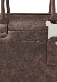 Braune Lederhandtasche mit strukturiertem Finish, Logo auf der Seite geprägt, dezente Nahtdetails und ein Metallschild, das am Griff befestigt ist.