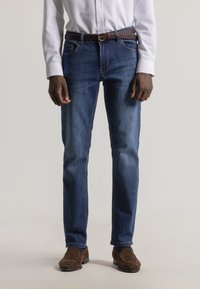 Jeans de mezclilla azul oscuro con corte recto, presentan un desgastado sutil y costuras en contraste. Combinados con un cinturón trenzado marrón.