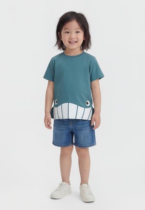 Χαμογελαστό παιδί με μεσαίου μήκους μαλλιά που φοράει μια τιρκουάζ μπλούζα με σχέδιο φάλαινας, τζιν σορτς και λευκά αθλητικά, στέκεται μπροστά σε λευκό φόντο.