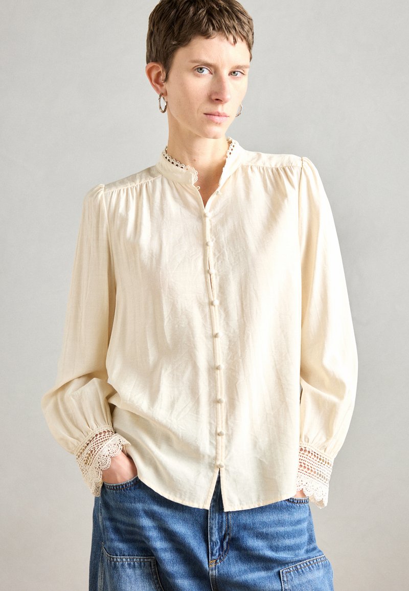 Blusa button-up color crema con colletto alto, dettagli in pizzo ai polsini e tessuto strutturato. Abbinata a denim.