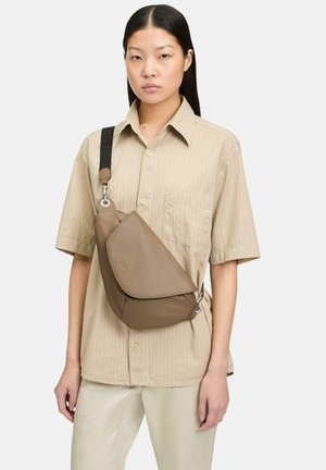 Braune Leder-Schultertasche mit geschwungenem Design, ausgestattet mit einem schwarzen verstellbaren Riemen und silbernen Beschlägen. Von einem Model als Crossbody-Tasche getragen.