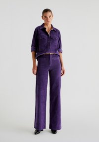 Set composto da una camicia in corduroy viola e pantaloni a gamba larga, caratterizzati da due tasche frontali e una finitura testurizzata, abbinato a scarpe nere a punta.