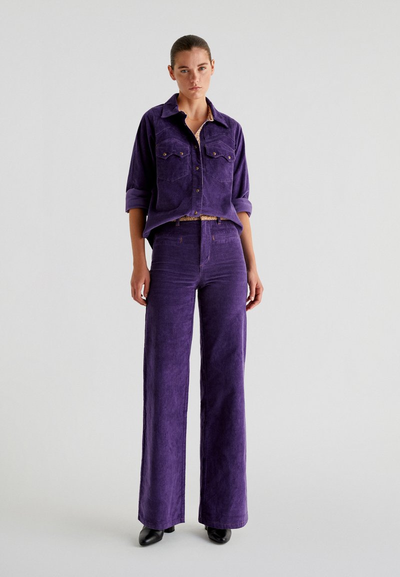 Set composto da una camicia in corduroy viola e pantaloni a gamba larga, caratterizzati da due tasche frontali e una finitura testurizzata, abbinato a scarpe nere a punta.