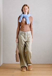 Haut à manches longues marron à côtes, écharpe bleu clair, pantalon large beige, chaussures couleur tan, tenant un sac marron, debout sur un sol en bois contre un mur uni.