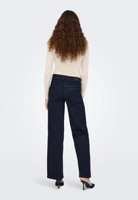 Femme aux longs cheveux bouclés portant un haut crème à manches longues, un jean foncé à jambes larges et des chaussures à talons noirs, debout de dos.