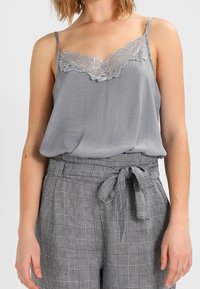 Camisola in satin grigio con pizzo, abbinata a pantaloni a quadri a vita alta con cintura. Entrambi i capi hanno una texture liscia.