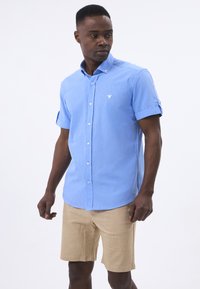 Kurzarm blaues Hemd mit Button-Down-Kragen, subtiler Textur und einem weißen gestickten Vogel-Logo, kombiniert mit beigen Shorts.