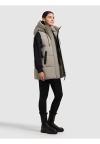 Bež puhuvest, millel on kõrge kaelus, tõmblukuga taskud ja vabalt langev lõige, kantud musta pikakäiselise ülikonna peale ning kombineeritud mustade retuuside ja saapatega.