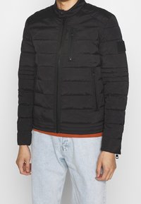 Black padded jacket with a stand-up collar, zippered pockets, and textured surface. Features ribbed hem and cuffs. Worn over an orange shirt.
