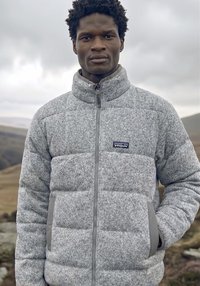 Muž ve světlé šedé fleecové bundě Patagonia s kapsami v rukou, stojící venku s zataženou oblohou a kopci v pozadí.