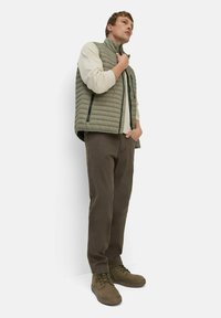Hellgrüne gesteppte Weste über einem cremefarbenen Langarmshirt, kombiniert mit dunkelbraunen Hosen und olivgrünen Schuhen, die ein einfaches Design und Texturen aufweisen.